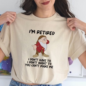 May include: A beige t-shirt with a cartoon image of a grumpy dwarf with a white beard and red shirt. The text "I'm Retired" is above the image and the text "I don't have to I don't want to You can't make me" is below the image.