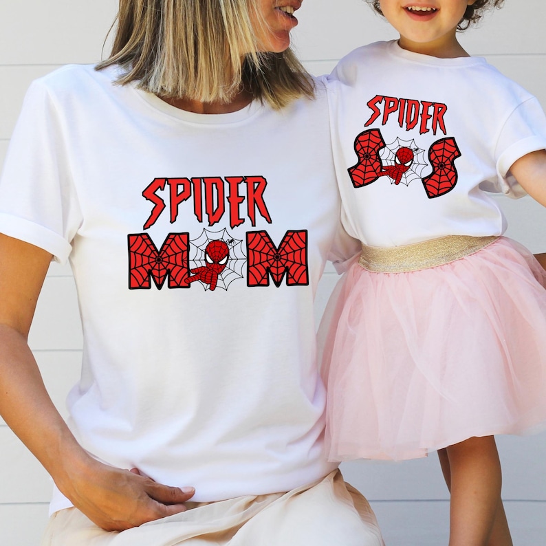 May include: A white t-shirt with the text "Spider Mom" in red and black letters with a spider web design. A child is wearing a white t-shirt with the text "Spider" in red and black letters with a spider web design.