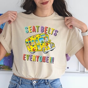 May include: A beige t-shirt with a yellow school bus graphic and the text "SEAT BELTS EVERYONE!!!" in rainbow colors.