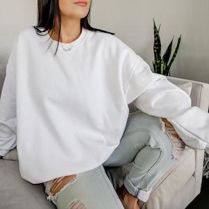 Peut inclure: Un sweat-shirt blanc surdimensionn&eacute; &agrave; manches longues et col rond. Le sweat-shirt est port&eacute; par une personne assise sur un canap&eacute; avec un jean bleu clair d&eacute;lav&eacute;.