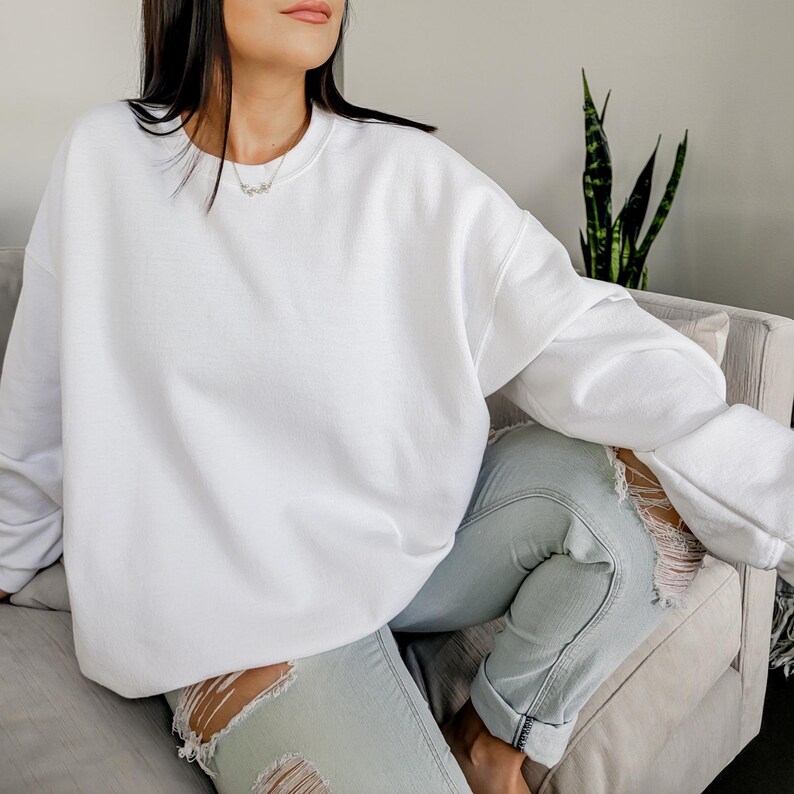Peut inclure: Un sweat-shirt blanc surdimensionn&eacute; &agrave; manches longues et col rond. Le sweat-shirt est port&eacute; par une personne assise sur un canap&eacute; avec un jean bleu clair d&eacute;lav&eacute;.