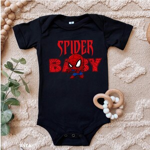 May include: Black baby bodysuit with the words "SPIDER BABY" in red, featuring a cartoon Spider-Man graphic. The bodysuit has short sleeves and three poppers at the bottom. A wooden teething ring with white beads is next to the bodysuit.