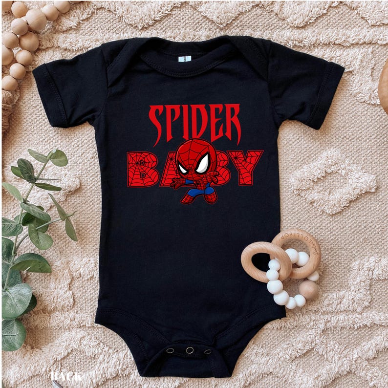 May include: Black baby bodysuit with the words "SPIDER BABY" in red, featuring a cartoon Spider-Man graphic. The bodysuit has short sleeves and three poppers at the bottom. A wooden teething ring with white beads is next to the bodysuit.