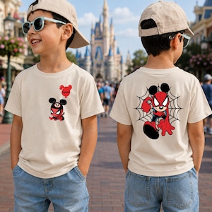 May include: A beige short-sleeved t-shirt featuring a small graphic on the front left chest of a cartoon character wearing a spider mask and holding a red balloon with the word Name, and a large graphic on the back of the character in a spider web.