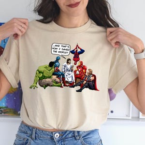 Puede incluir: Camiseta beige con un gr&aacute;fico de superh&eacute;roes y Jesucristo. La imagen incluye el texto: "...&iexcl;Y AS&Iacute; ES COMO SALV&Eacute; EL MUNDO!" Se representan a Hulk, Capit&aacute;n Am&eacute;rica, Iron Man, Thor y Spider-Man.