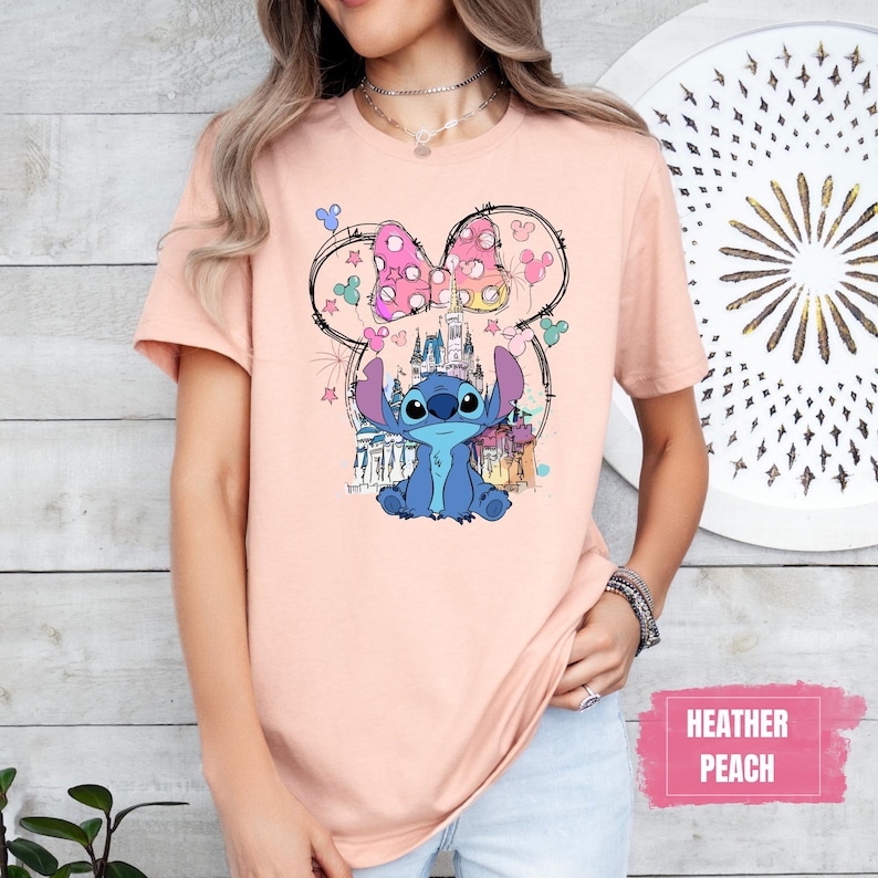 May include: Heather peach t-shirt featuring a colorful graphic of the character Stitch from Lilo & Stitch, with a Minnie Mouse bow and castle. The design includes stars and balloons. The text "Heather Peach" is in a pink rectangle.