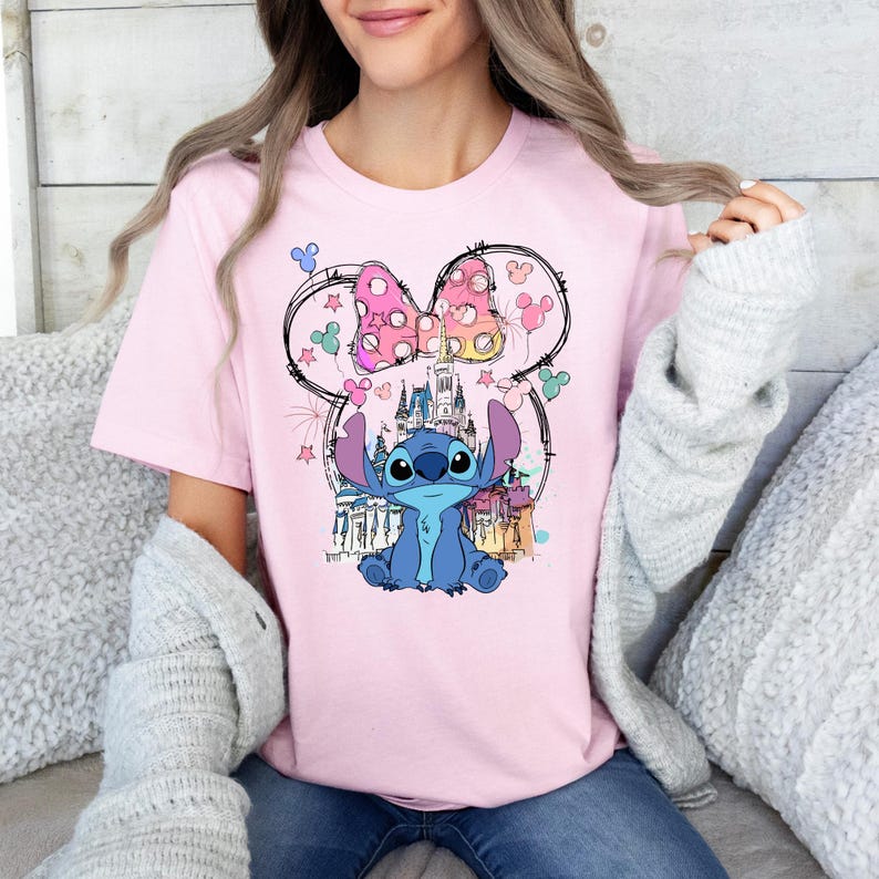 May include: A light pink t-shirt featuring a graphic of the character Stitch from Lilo & Stitch. The design includes a castle, a bow, and other decorative elements. The shirt is a crew neck style.