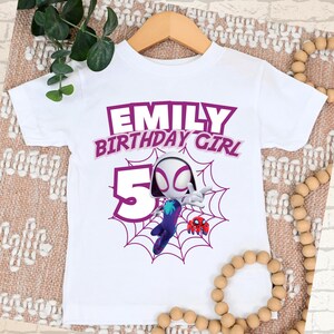 Pu&ograve; includere: T-shirt bianco con grafica viola che recita "EMILY BIRTHDAY GIRL 5". La maglietta presenta un personaggio dei cartoni animati in un disegno a ragnatela. La maglietta &egrave; esposta su una gruccia di legno.