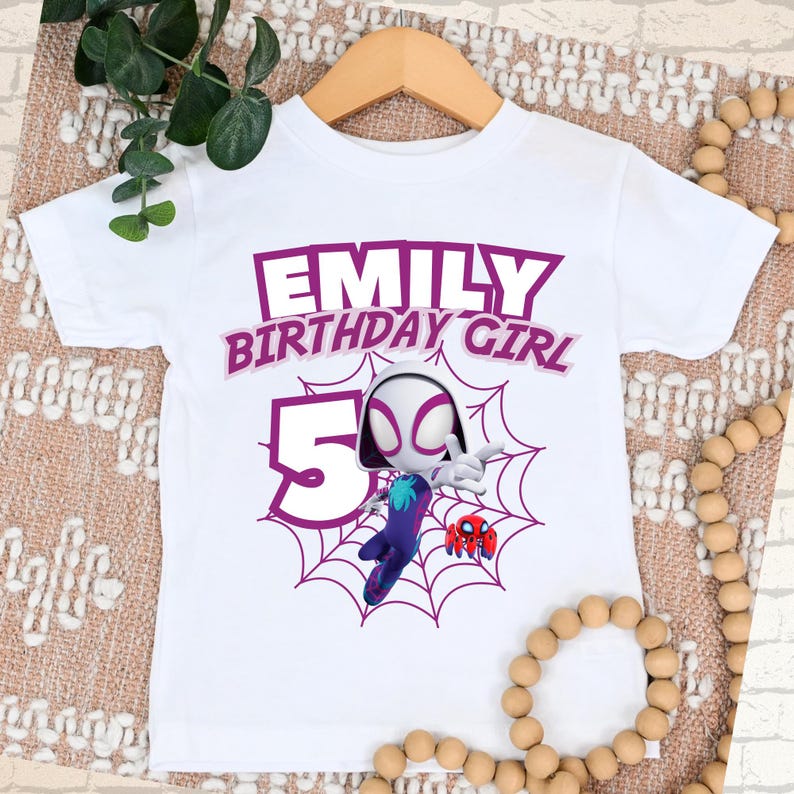 Pu&ograve; includere: T-shirt bianco con grafica viola che recita "EMILY BIRTHDAY GIRL 5". La maglietta presenta un personaggio dei cartoni animati in un disegno a ragnatela. La maglietta &egrave; esposta su una gruccia di legno.