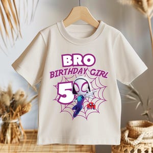 Pu&ograve; includere: T-shirt color crema a maniche corte con grafica di un supereroe e la scritta "BRO BIRTHDAY GIRL 5". Il design include una ragnatela e un personaggio dei cartoni animati viola e blu.