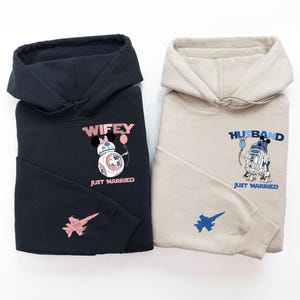 Pode incluir: Duas sweatshirts com capuz pretas e bege com um design tem&aacute;tico de Star Wars. A sweatshirt preta tem um BB-8 rosa e branco com um la&ccedil;o rosa e um bal&atilde;o rosa. A sweatshirt bege tem um R2-D2 azul e branco com um bal&atilde;o azul. Ambas as sweatshirts dizem "Just Married" e t&ecirc;m um pequeno gr&aacute;fico de avi&atilde;o na manga.