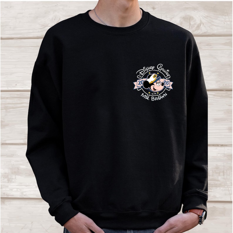 Op de afbeelding: Zwarte crewneck sweatshirt met een Disney Cruise ontwerp. Het ontwerp bevat Mickey Mouse met een kapiteinshoed, de woorden "Disney Cruise", het jaar "26" en de naam "Mr. Brown".