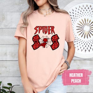 May include: A peach colored t-shirt with a red spider-man graphic and the text "SPIDER" in black letters. The shirt is made of a soft, comfortable material.