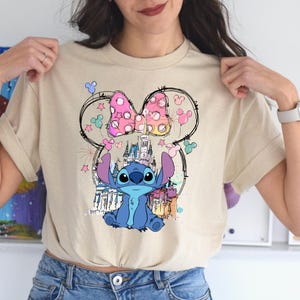 May include: Beige t-shirt featuring a colorful graphic of the character Stitch from Lilo & Stitch. The design includes a pink polka-dot bow, a castle, and Mickey Mouse silhouettes. The shirt is a casual wear item.