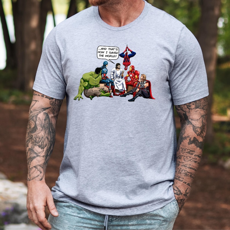 Puede incluir: Camiseta gris con una ilustraci&oacute;n estilo c&oacute;mic de Jes&uacute;s y superh&eacute;roes. La imagen incluye a Hulk, Capit&aacute;n Am&eacute;rica, Iron Man, Thor y Spider-Man. El texto en un bocadillo dice: "...&iexcl;Y AS&Iacute; ES COMO SALV&Eacute; EL MUNDO!"