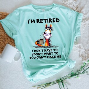 Pu&ograve; includere: Una maglietta turchese con il testo "I'm Retired" e un cavallo dei cartoni animati che tiene una tazza di caff&egrave;. Il testo sotto il cavallo dice "I don't have to I don't want to You can't make me."