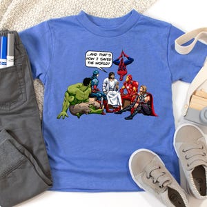 May include: A light blue t-shirt featuring a cartoon illustration of Jesus Christ with several Marvel superheroes. The text in a speech bubble reads, "...AND THAT'S HOW I SAVED THE WORLD!" The shirt is paired with gray pants and gray sneakers.