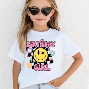 May include: White t-shirt with the words "BIRTHDAY GIRL" in pink and black checkered design. The shirt features a yellow smiley face with a star, flowers, and other graphics. The shirt is worn by a person wearing heart-shaped sunglasses.