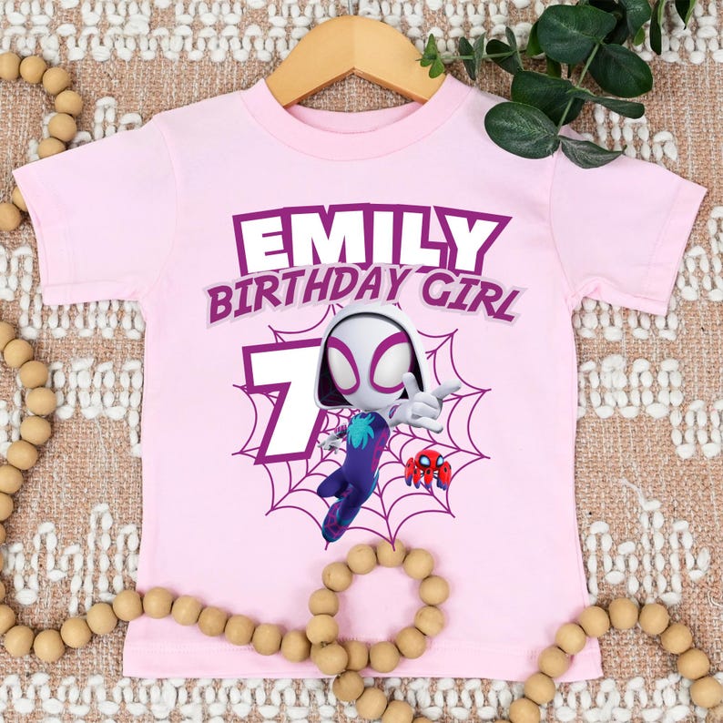 Pu&ograve; includere: Una maglietta rosa chiaro con la scritta "EMILY BIRTHDAY GIRL" in viola. Sono presenti anche il numero 7 e un personaggio di Spider-Gwen. La maglietta &egrave; appesa a una gruccia di legno.
