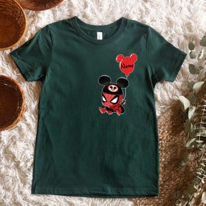 May include: A dark green short-sleeve t-shirt featuring a graphic of a character wearing a red and black spider-themed mask with mouse ears. The character holds a red balloon shaped like mouse ears with the word Name printed in black text.