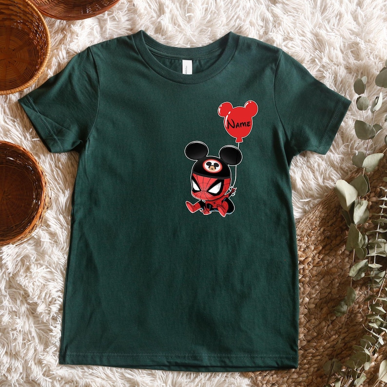 May include: A dark green short-sleeve t-shirt featuring a graphic of a character wearing a red and black spider-themed mask with mouse ears. The character holds a red balloon shaped like mouse ears with the word Name printed in black text.
