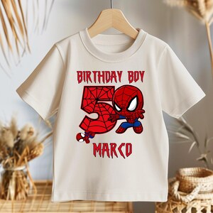 May include: Cream-coloured t-shirt with a Spider-Man graphic. The design includes the text "BIRTHDAY BOY 5 MARCO" in red, with a large number 5 and a Spider-Man character illustration. The shirt is hanging on a wooden hanger.