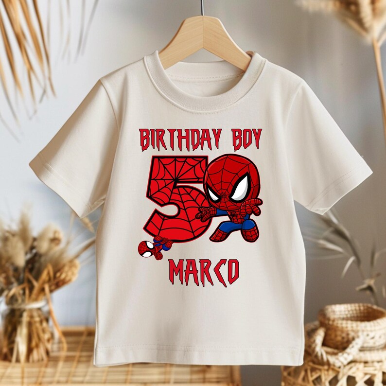 May include: Cream-coloured t-shirt with a Spider-Man graphic. The design includes the text "BIRTHDAY BOY 5 MARCO" in red, with a large number 5 and a Spider-Man character illustration. The shirt is hanging on a wooden hanger.