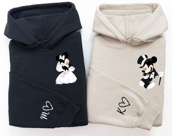 May include: A pair of black and beige hoodies with a Mickey Mouse and Minnie Mouse wedding design. The black hoodie has Minnie Mouse in a wedding dress and the beige hoodie has Mickey Mouse in a tuxedo. The hoodies have the initials "M" and "K" with a heart on the sleeves.