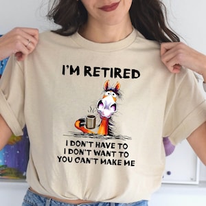 Pu&ograve; includere: Una maglietta beige con un cavallo dei cartoni animati che tiene una tazza di caff&egrave;. Il testo sulla maglietta recita "I'm Retired I don't have to I don't want to You can't make me."