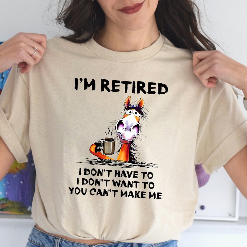 Pu&ograve; includere: Una maglietta beige con un cavallo dei cartoni animati che tiene una tazza di caff&egrave;. Il testo sulla maglietta recita "I'm Retired I don't have to I don't want to You can't make me."