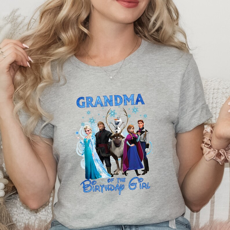 May include: A grey crew neck t-shirt with the word "GRANDMA" in blue, and a graphic of characters from the film Frozen. Below the graphic, the text reads "OF THE BIRTHDAY GIRL" in blue. The shirt is a crew neck.