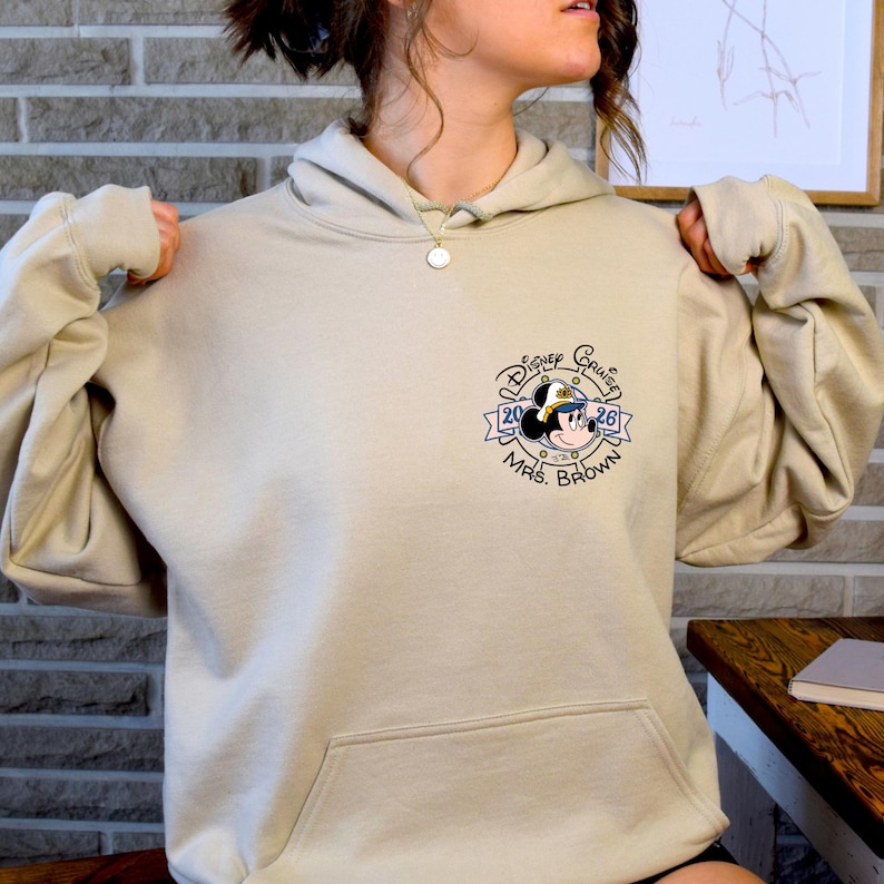 Op de afbeelding: Een beige hoodie met een voorzak en een trekkoord. De linkerbovenborst toont een Disney Cruise-afbeelding met Mickey Mouse met een kapiteinshoed en de tekst "Disney Cruise 2026 Mrs. Brown."