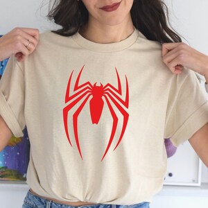 Op de afbeelding: Een cr&egrave;mekleurig T-shirt met een rood Spiderman-logo op de voorkant.