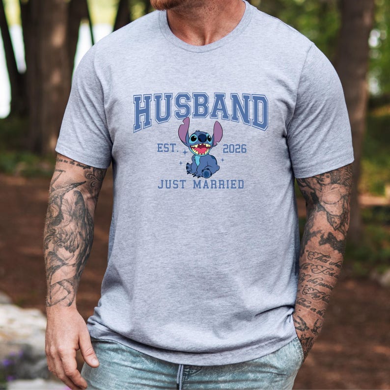 Op de afbeelding: Lichtgrijs t-shirt met het woord "HUSBAND" in blauwe blokletters boven een stripfiguur en de tekst "EST. 2026 JUST MARRIED". De stripfiguur is blauw met grote oren en een brede glimlach.