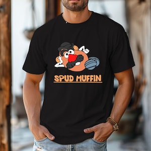 May include: Black t-shirt featuring a cartoon potato character with a black hat, red nose, and a blue ball. The text "SPUD MUFFIN" is printed in orange below the graphic. The shirt is made of a soft material.
