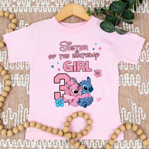 以下が含まれることがあります： 「Sister of the Birthday Girl」の文字と数字「3」がプリントされた、薄いピンク色のTシャツ。2人の漫画のキャラクターが抱き合っているグラフィックが特徴です。Tシャツは丸首で半袖です。
