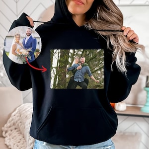 May include: Black hoodie with a photo printed on the front. The photo shows a couple in a forest setting. The woman is wearing a white dress and the man is wearing a blue shirt.