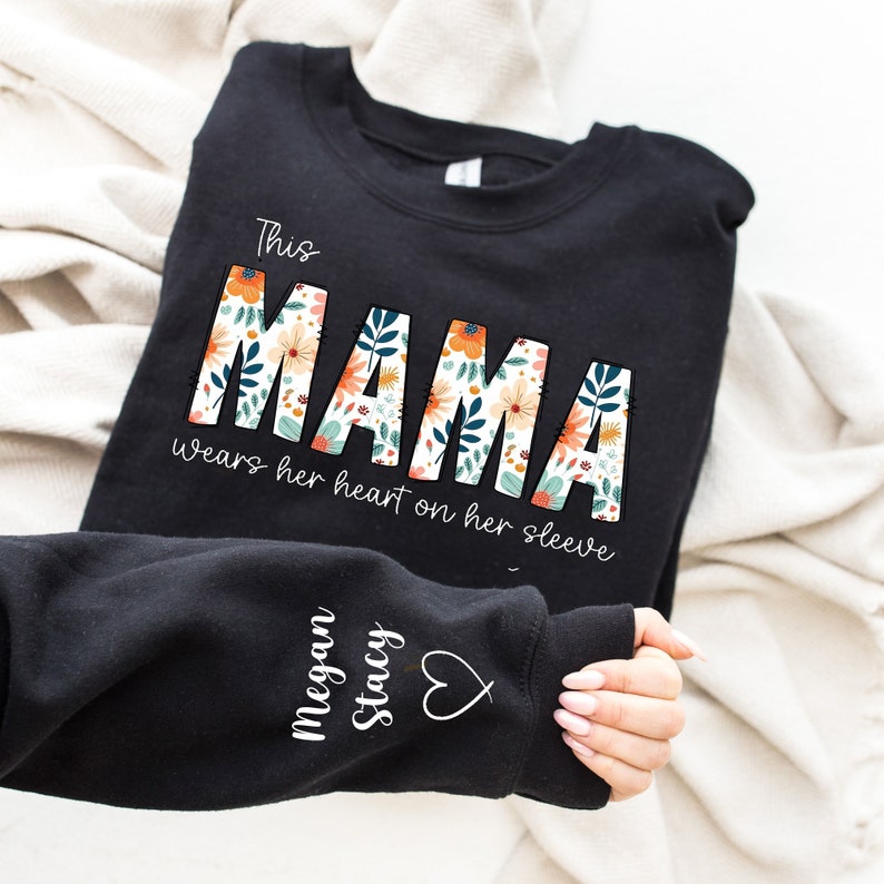 May include: Black sweatshirt with the text "This MAMA wears her heart on her sleeve." The word "MAMA" is decorated with a floral design in orange, white, and blue. The sleeve features the name "Megan Stacy" and a heart symbol.