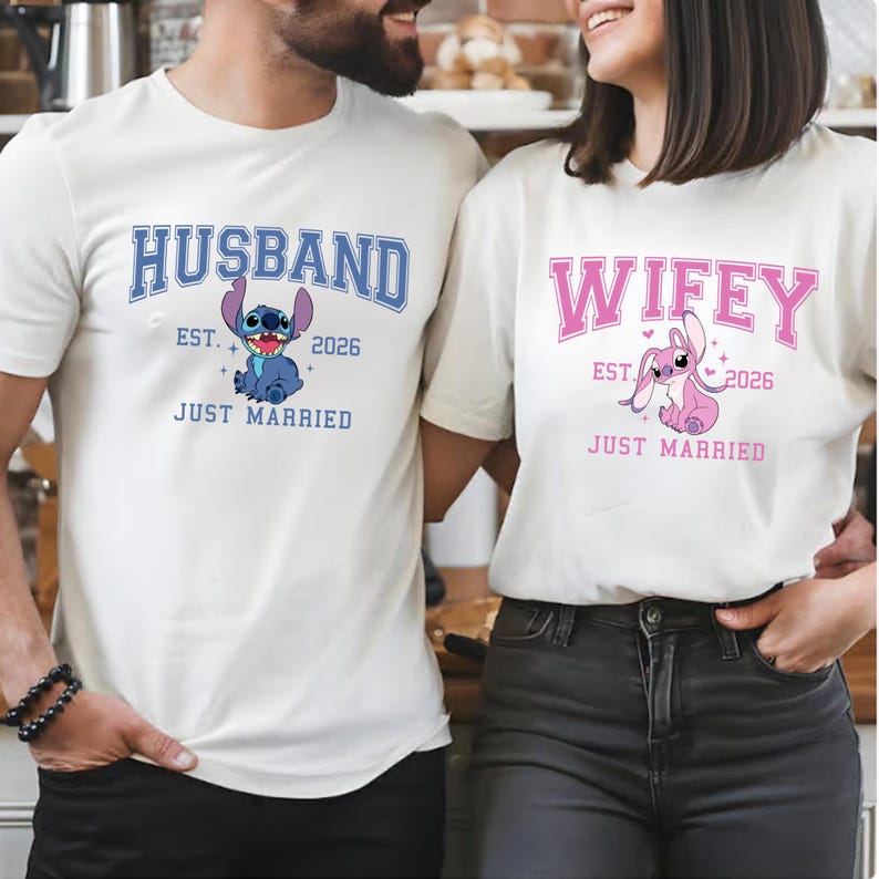 Op de afbeelding: Cr&egrave;mekleurige T-shirts met bijpassende ontwerpen. E&eacute;n shirt toont het woord "HUSBAND" in blauw, een cartoonfiguur en "EST. 2026 JUST MARRIED". De andere zegt "WIFEY" in roze, met een cartoonfiguur en "EST. 2026 JUST MARRIED".