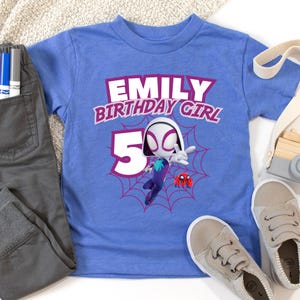 Pu&ograve; includere: Una maglietta azzurra con la scritta "EMILY BIRTHDAY GIRL 5" e una grafica a cartoni animati di un supereroe. La maglietta &egrave; abbinata a pantaloni grigi e scarpe da ginnastica grigio chiaro.