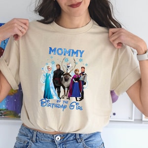 May include: Beige t-shirt with a Frozen movie graphic, including Elsa, Anna, Kristoff, and Olaf. The text "MOMMY of the BIRTHDAY GIRL" is printed in blue above the characters. The shirt has short sleeves and a crew neck.