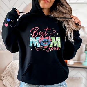 May include: Black hooded sweatshirt with the words "Best Mom Ever" in pink and green, featuring a cartoon character. The left sleeve has the names "Oliver" and "Katie" with heart icons. A cozy and stylish gift.