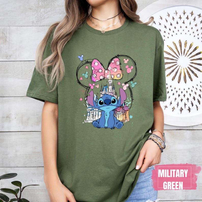 May include: Olive green t-shirt featuring a cartoon Stitch character with a pink bow and castle design. The design is set within a Mickey Mouse ear outline. The text "MILITARY GREEN" is printed on a pink rectangle.