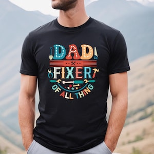 May include: Black t-shirt with a colorful graphic that says "Dad Fixer of All Thing" with various tools and stars around the text.