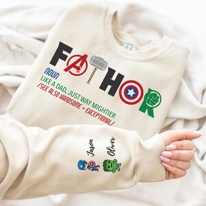 May include: Beige sweatshirt with the word "FATHOR" in black and red, featuring superhero symbols. The sleeve displays the names "Jason" and "Oliver" with cartoon superhero characters. The text reads "NOUN LIKE A DAD, JUST WAY MIGHTIER /SEE ALSO HANDSOME + EXCEPTIONAL/."