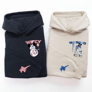 Pode incluir: Duas sweatshirts com capuz, uma preta e outra bege, com um gr&aacute;fico tem&aacute;tico de Star Wars. A sweatshirt preta tem um BB-8 rosa e azul com a palavra "WIFEY" acima. A sweatshirt bege tem um R2-D2 azul e branco com a palavra "HUSBAND" acima.