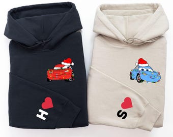 Puede incluir: Dos sudaderas con capucha dobladas. Una es negra con un diseño de coche rojo con gorro de Papá Noel y luces navideñas. La otra es beige con un diseño de coche azul con gorro de Papá Noel. Cada manga izquierda tiene un corazón y una letra.