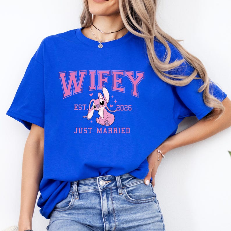 Op de afbeelding: Koningsblauw T-shirt met het woord "Wifey" in roze, boven "Est. 2026" en "Just Married". Een roze cartoonfiguur staat in het midden. Het shirt is een casual stijl.