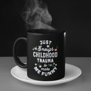 Puede incluir: Taza de caf&eacute; negra con un dise&ntilde;o blanco que dice "Just Enough Childhood Trauma to Make Me Funny" con peque&ntilde;os detalles florales.