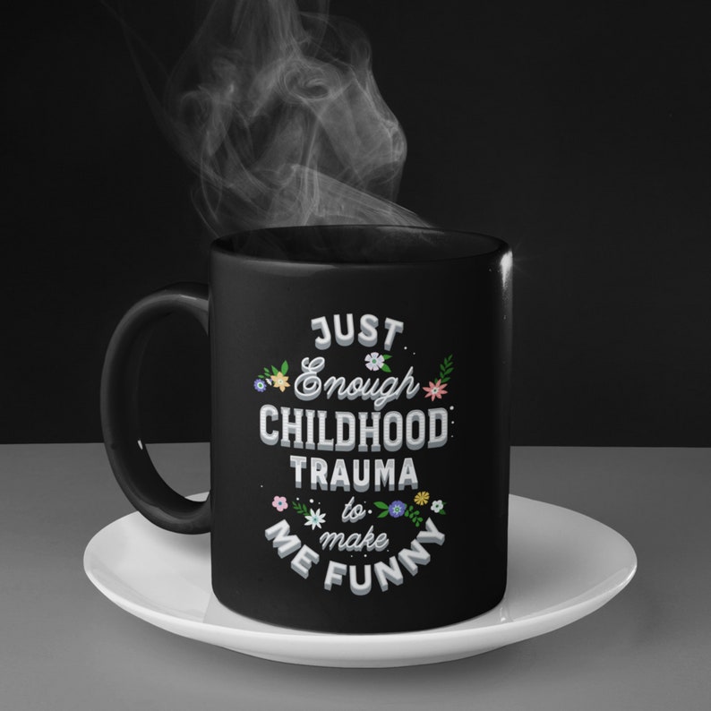 Puede incluir: Taza de caf&eacute; negra con un dise&ntilde;o blanco que dice "Just Enough Childhood Trauma to Make Me Funny" con peque&ntilde;os detalles florales.